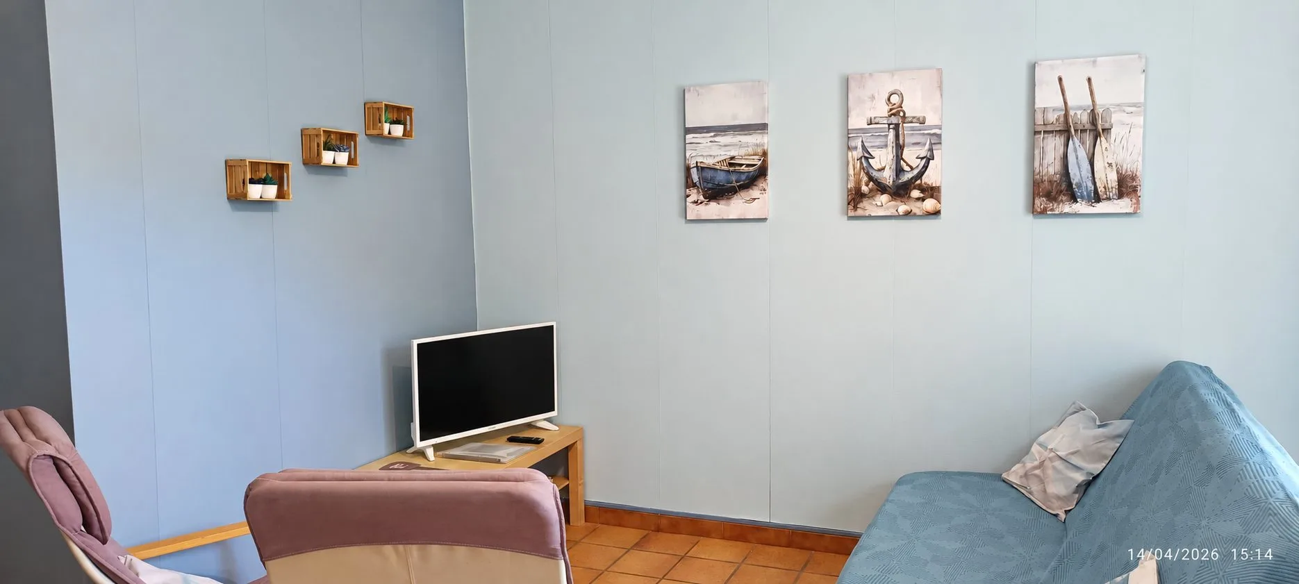 Un salon aux murs bleu clair décoré de trois tableaux marins représentant une barque, une ancre et des rames, avec une télévision, un canapé et des fauteuils.