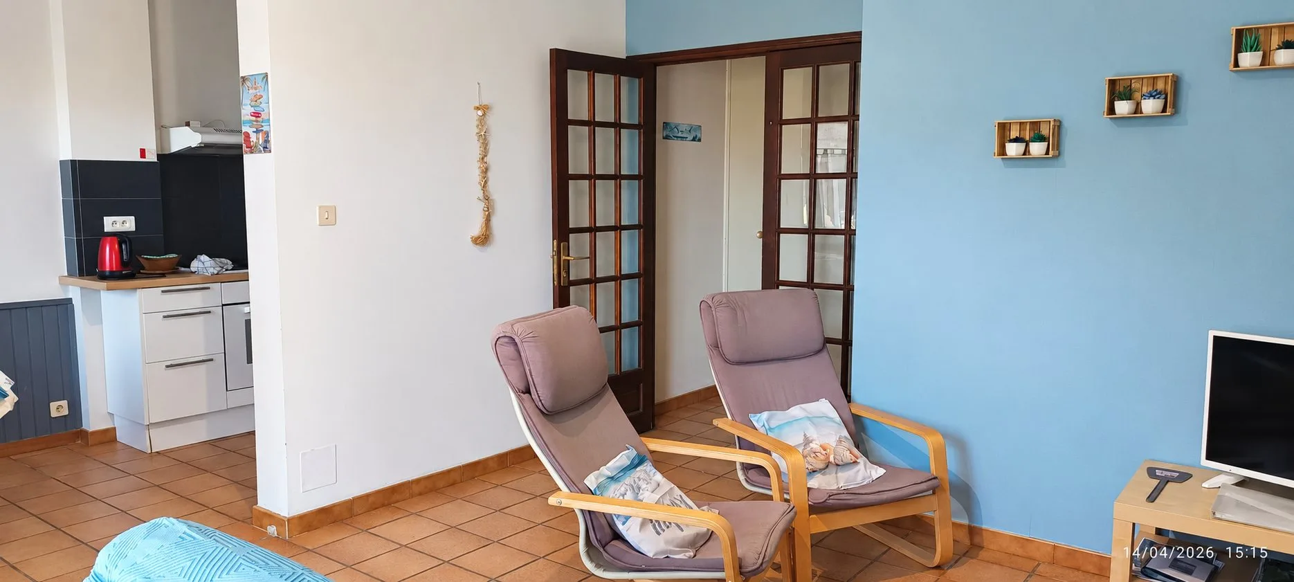 Pièce de vie avec deux fauteuils beige rosé, une porte vitrée en bois, des étagères murales bleues et une kitchenette au fond.