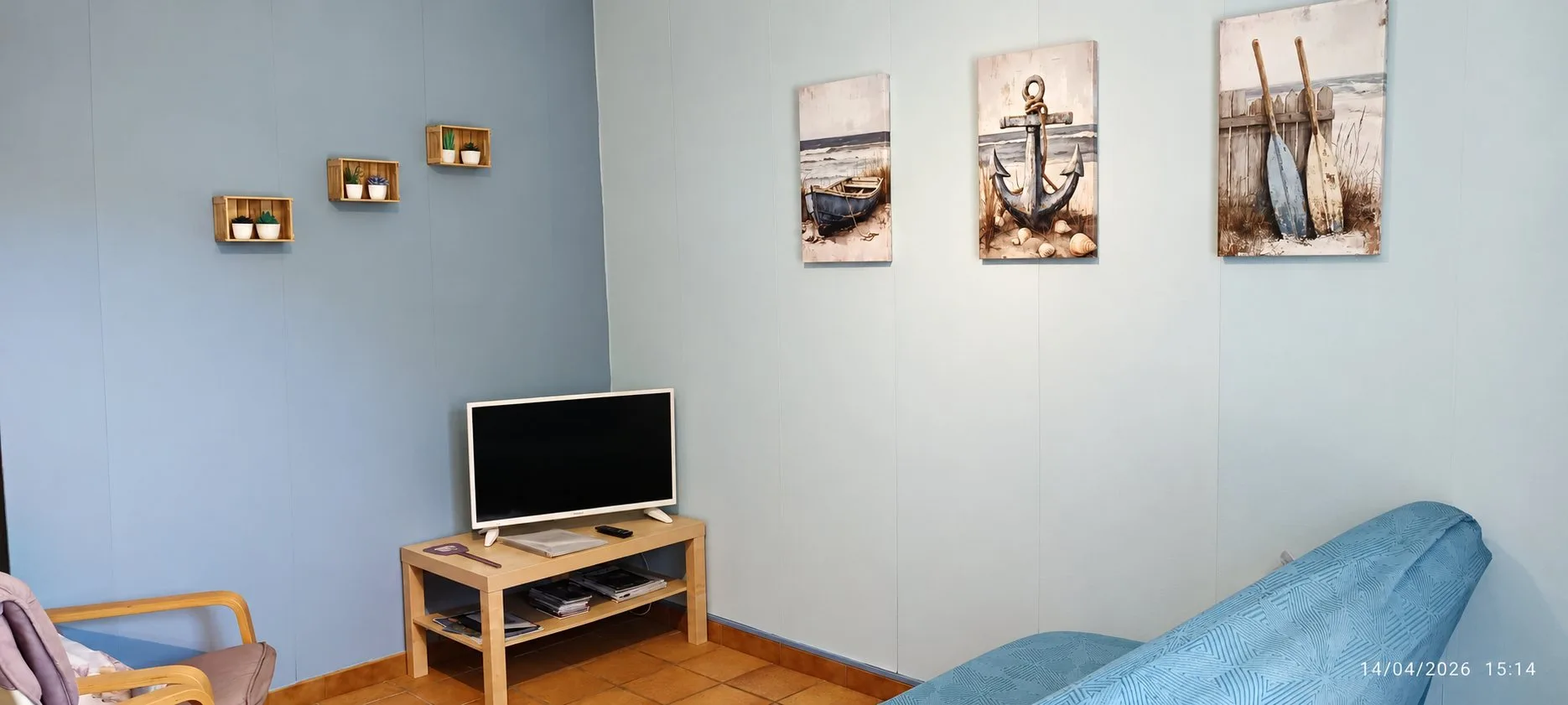 Un coin salon aux murs bleu-gris avec une télévision sur un meuble en bois clair, trois tableaux sur le thème marin, des petites étagères en cageots et un canapé bleu.