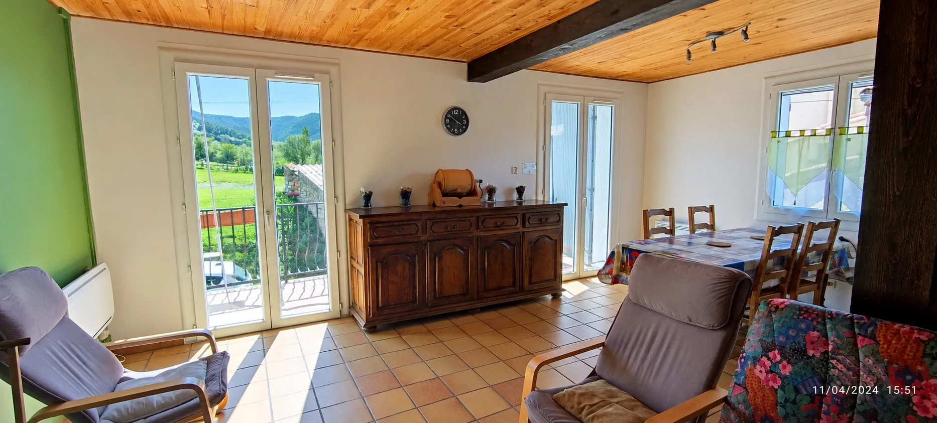 Pièce de vie avec buffet en bois, table et chaises, fauteuils et portes-fenêtres ouvrant sur un balcon et un paysage de montagne.