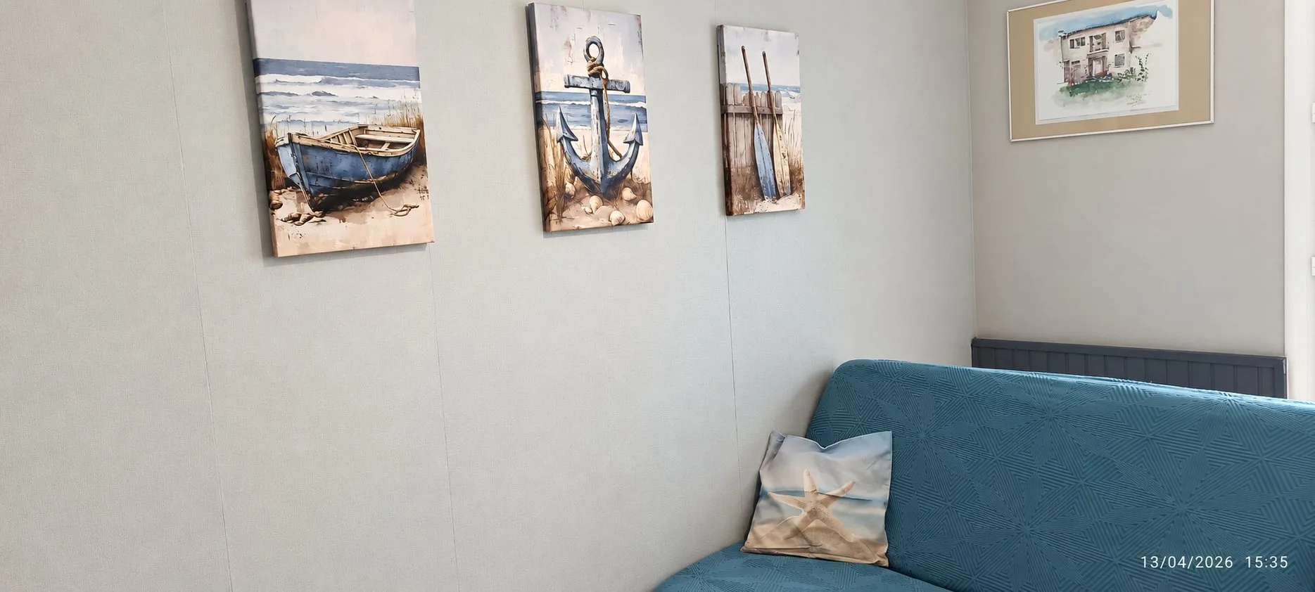 Un coin de pièce avec trois toiles sur le thème de la mer, un petit tableau encadré et un canapé bleu garni d'un coussin.