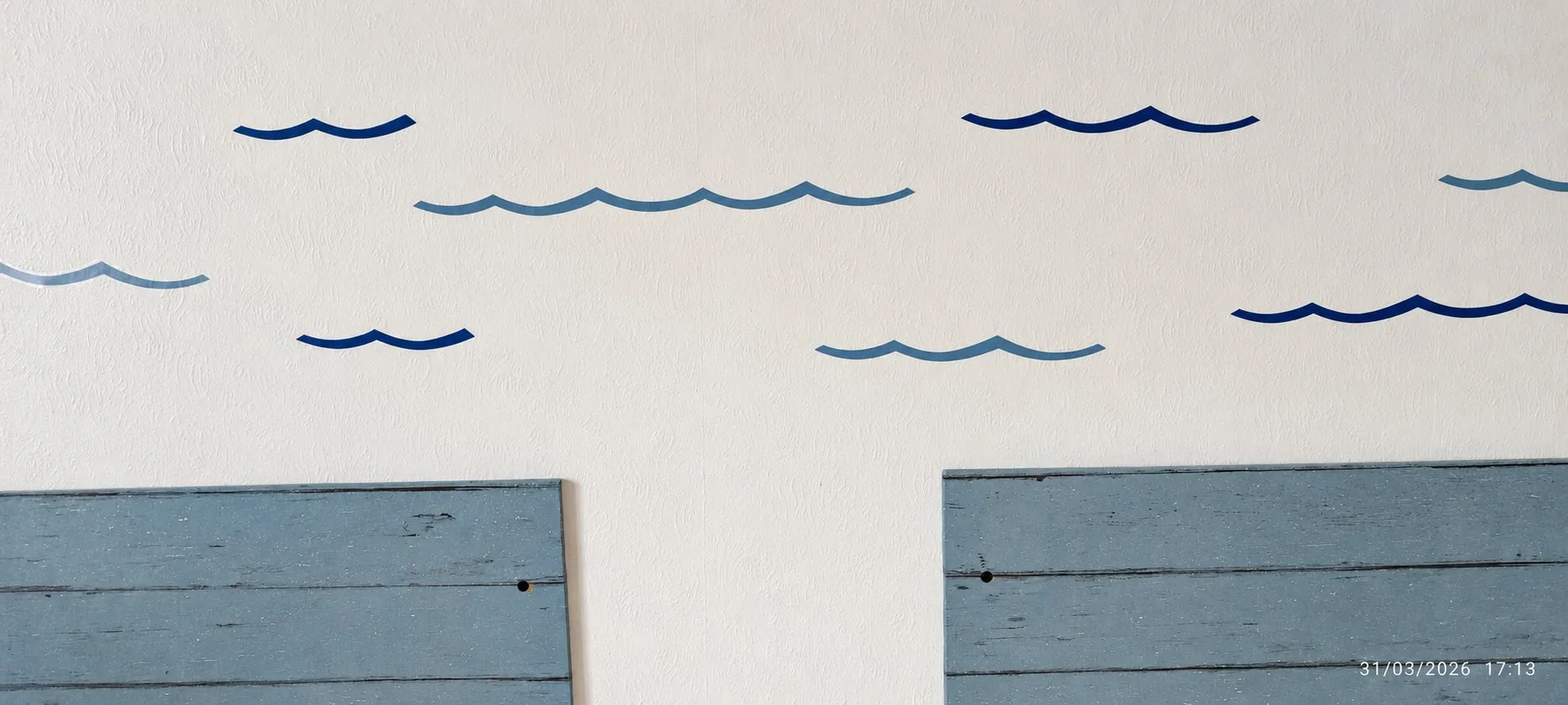 Des stickers en forme de vagues dans différents tons de bleu décorent un mur blanc au-dessus de deux panneaux en bois peint bleu-gris.