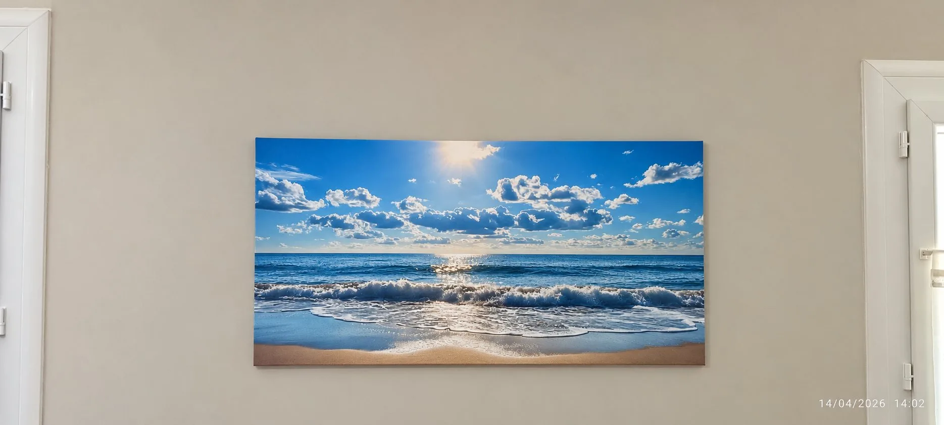 Un grand tableau représentant une plage, des vagues et un ciel bleu nuageux est accroché à un mur clair entre deux fenêtres.