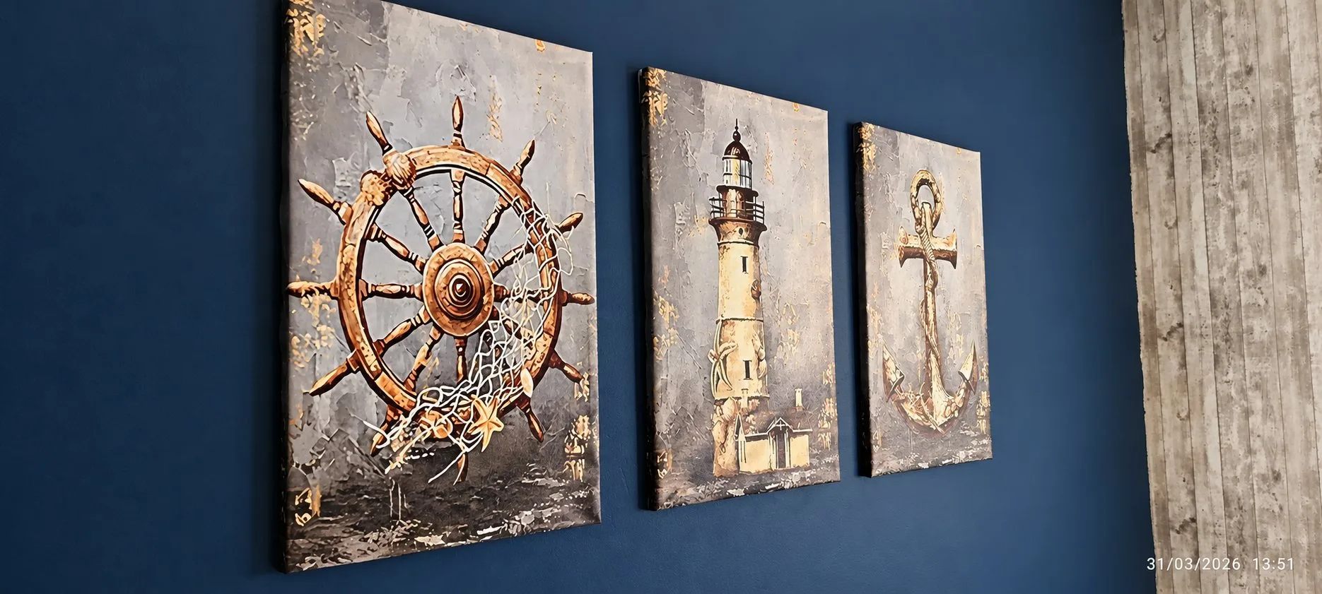 Trois toiles à thème marin accrochées sur un mur bleu marine représentent une barre de bateau avec filet, un phare et une ancre.