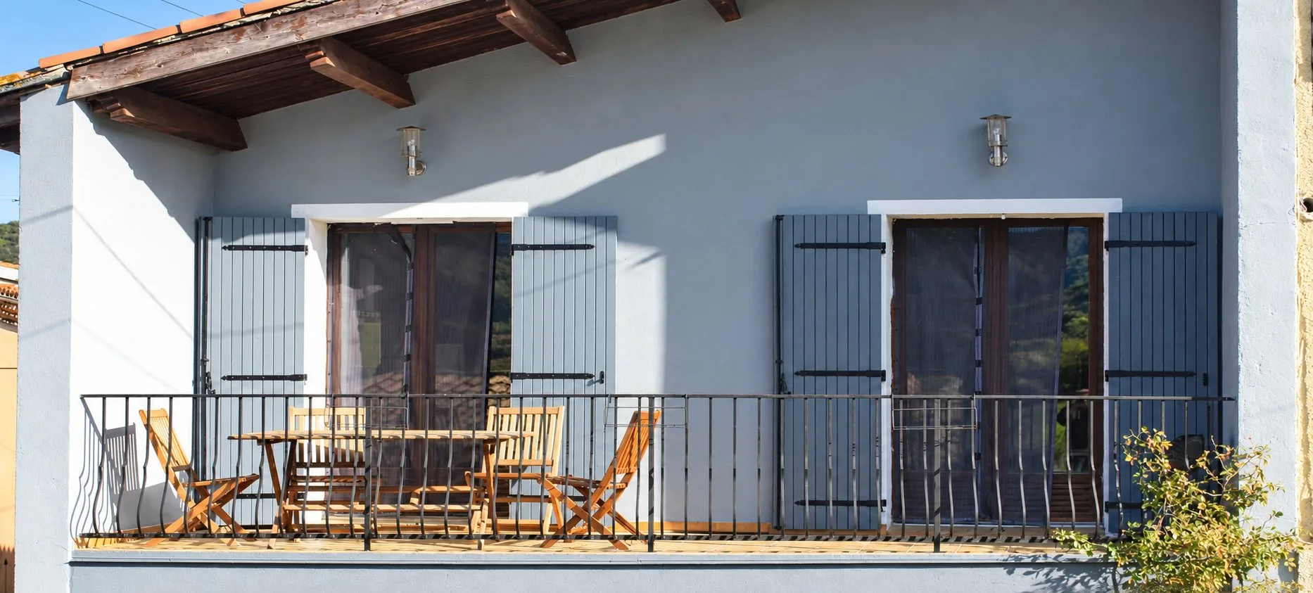 Une terrasse avec garde-corps métallique, table et chaises pliantes en bois devant une façade grise aux volets gris ouverts.
