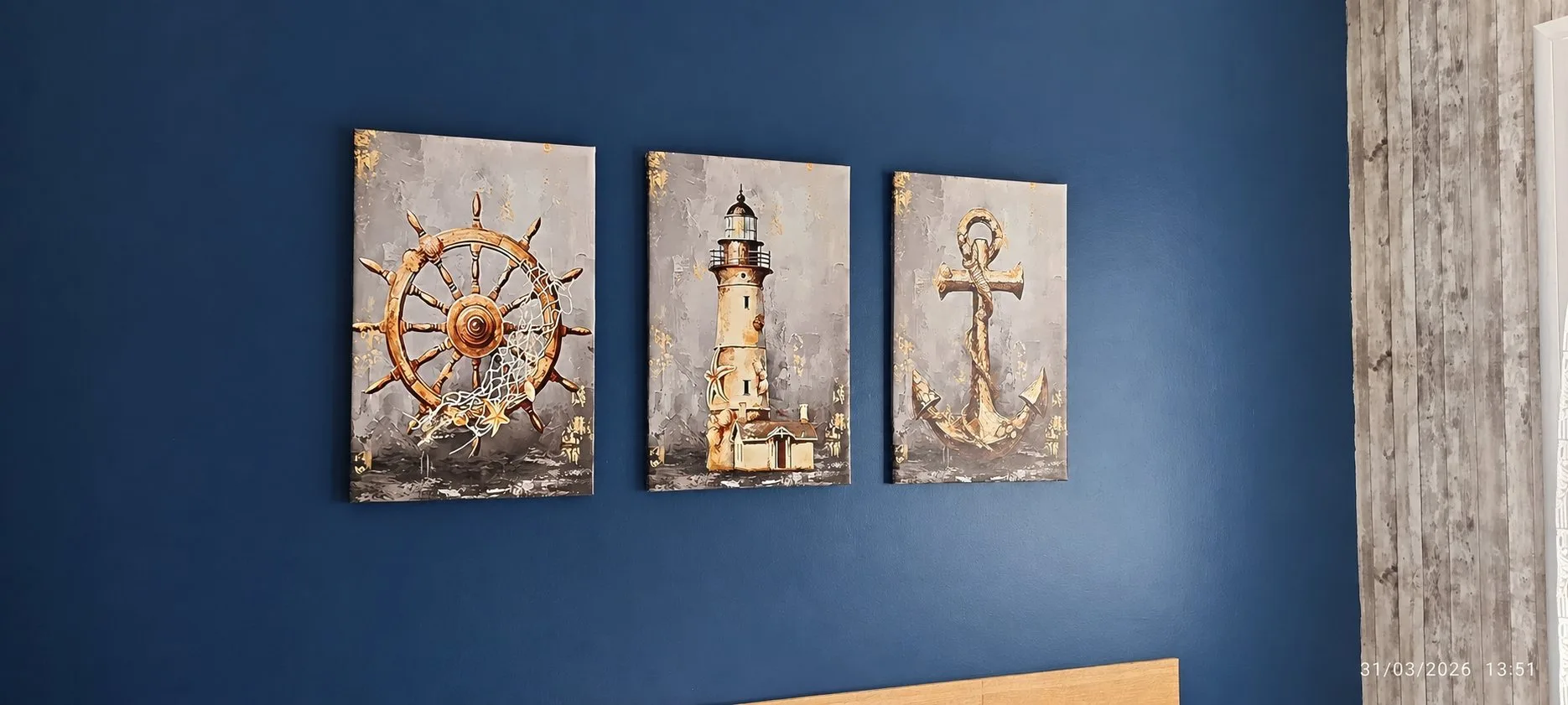 Trois tableaux représentant une barre de navire, un phare et une ancre sont accrochés côte à côte sur un mur bleu marine.