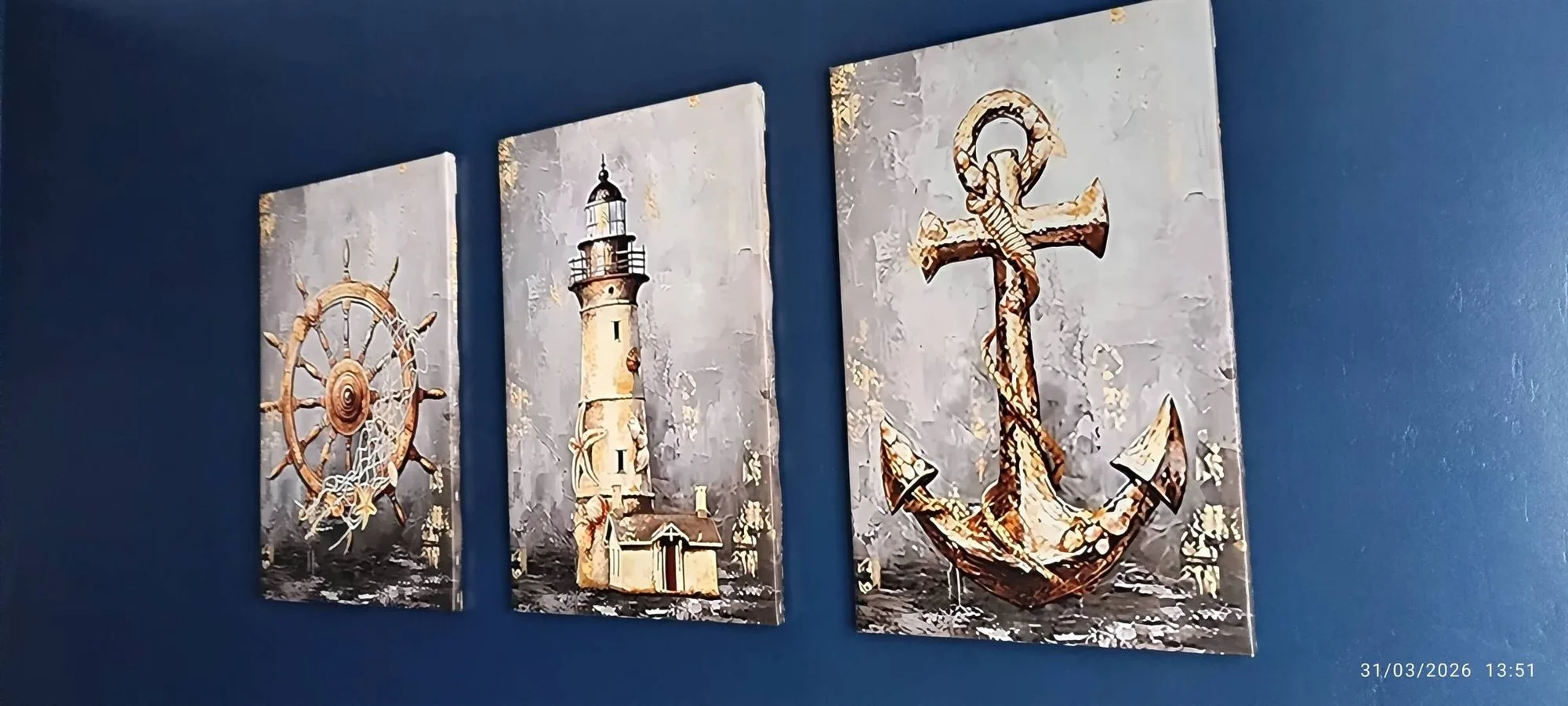 Trois toiles décoratives accrochées sur un mur bleu marine représentent une barre à roue de bateau, un phare et une ancre sur fond gris.